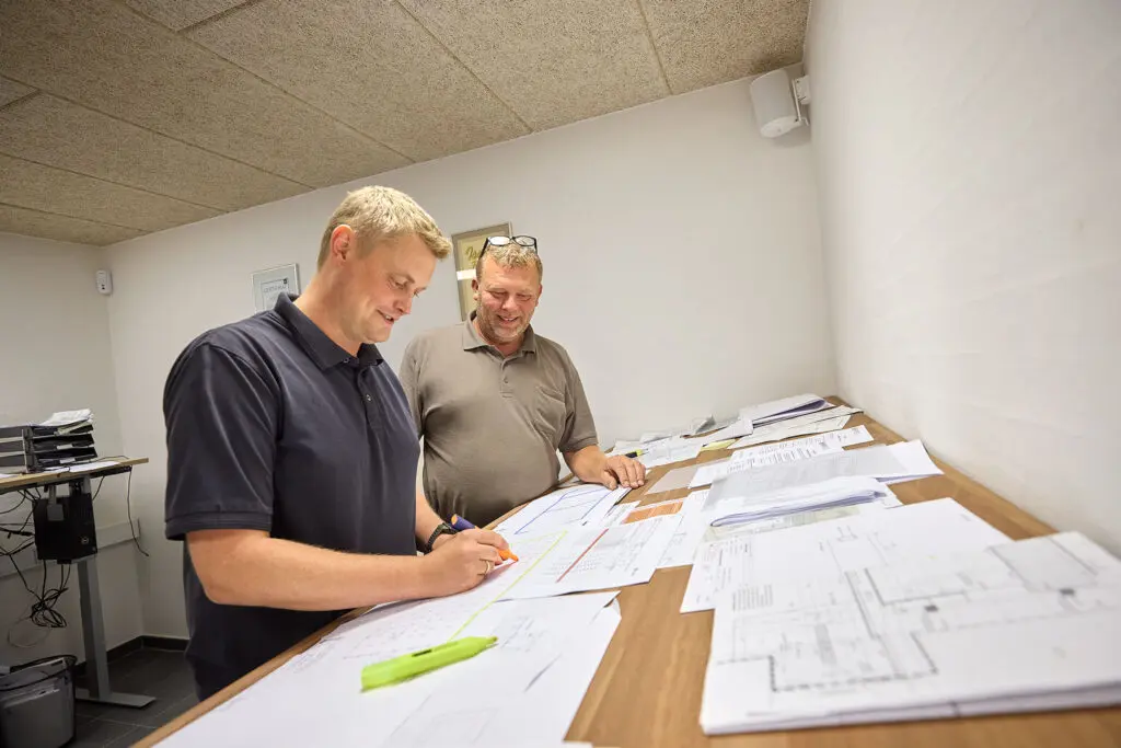 To mænd gennemgår og markerer byggetegninger ved et bord på et kontor.